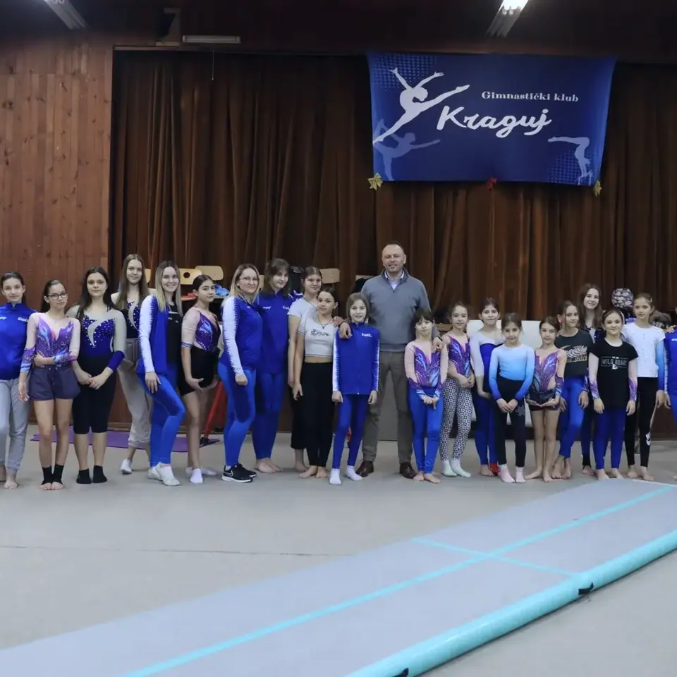 Članice kluba u trikoima i trenerkama stoje u redu ispod plavog klupskog banera „Gimnastički klub Kraguj“